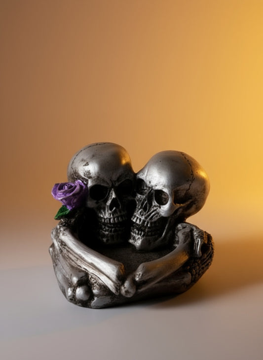 Decorative figurine of two skulls with a purple flower on a dark surface