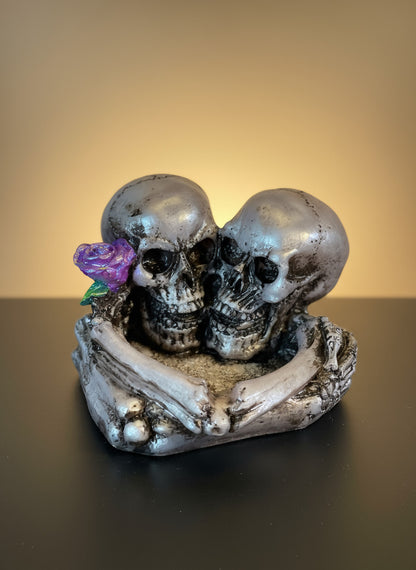 Two skulls embracing with a heart and purple flower on a dark background